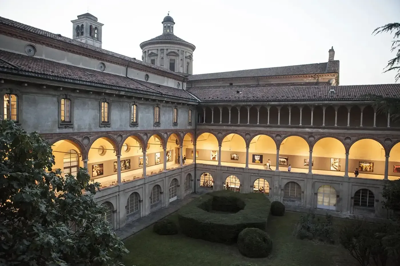 The museum's cloister photographed at dusk, a two-storey Renaissance courtyard with arched loggias lit from within, photographs hung along the upper arcade, and clipped box hedges in the garden below