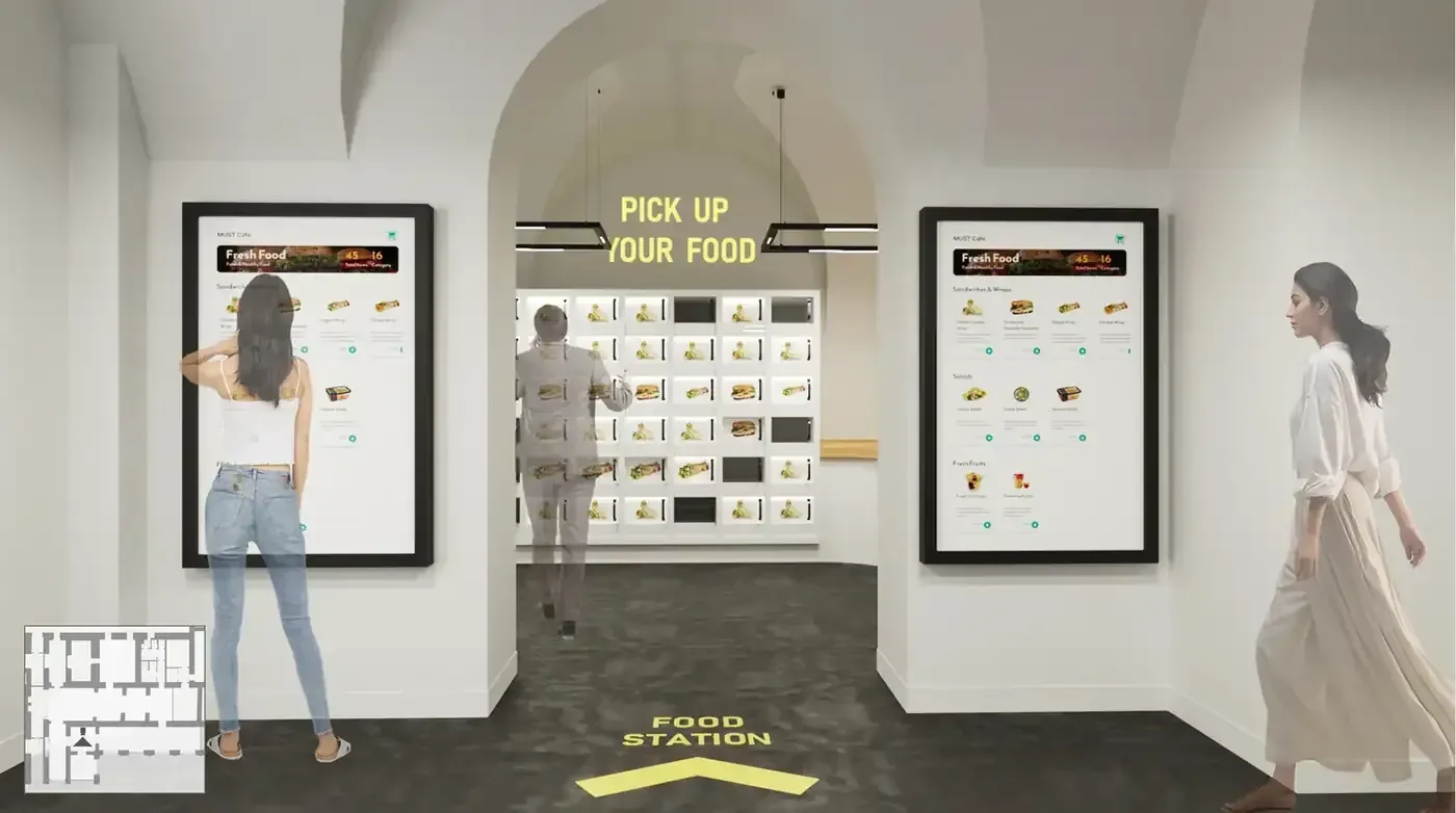 A render of the proposed food station, with two large wall-mounted touchscreens showing a MUST Cafe menu of sandwiches, wraps, salads and fresh fruit, a wall of illuminated food lockers behind an archway, the words pick up your food in yellow overhead and food station with an arrow painted on the floor