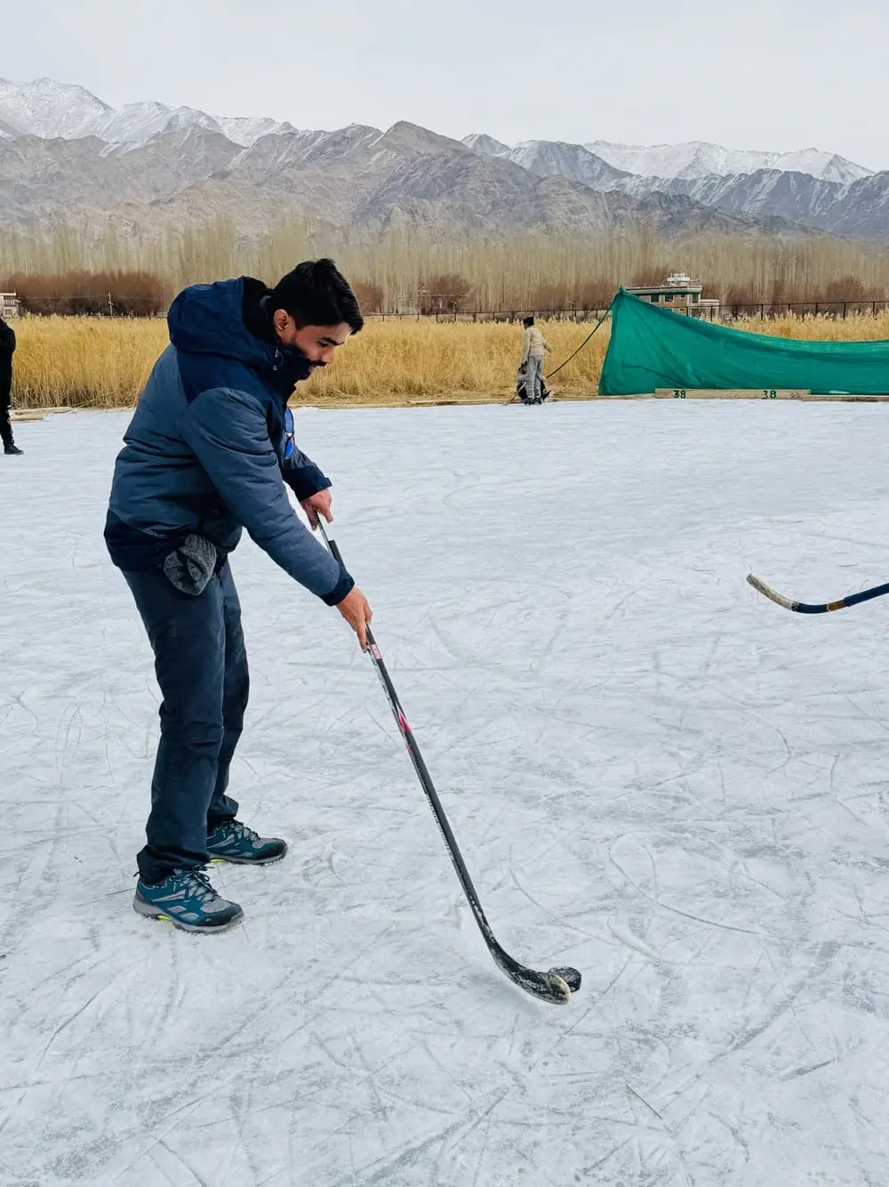 Gaus mid-swing with a hockey stick on an outdoor ice rink in Ladakh, snow-dusted mountains behind him.